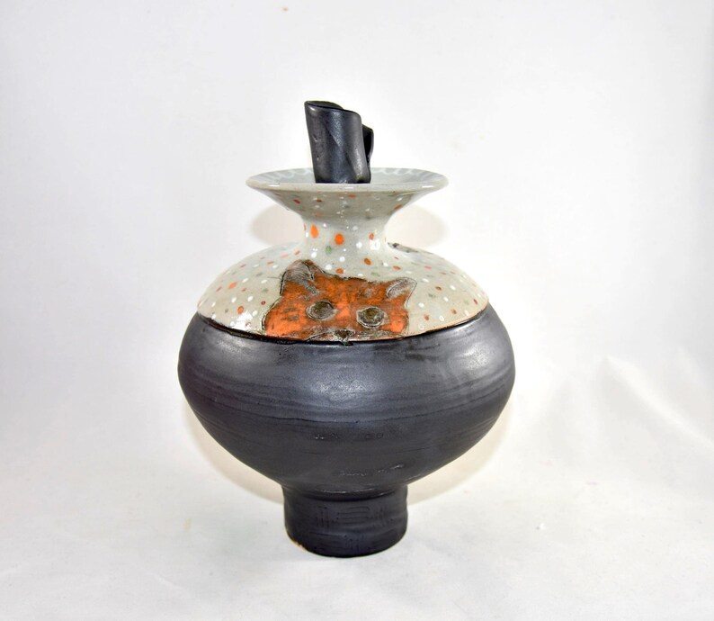 May include: A ceramic pet memorial urn with a black base and a light gray top decorated with orange polka dots. The lid features an orange cat face. A small black stopper fits into the top of this urn. The jar is a unique piece of art.