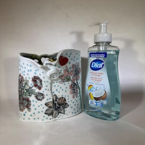 May include: A ceramic vase with a floral design and blue polka dots, next to a bottle of Dial coconut water and mango hand soap. The vase has a unique shape and a white rose accent.