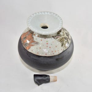 This is a handmade urn for holding a cat's ashes. It is ceramic and was made in the artist's Oregon pottery studio. This is a top view where you can see drawings of cats on the shoulder of the urn and the stopper laying beside it.