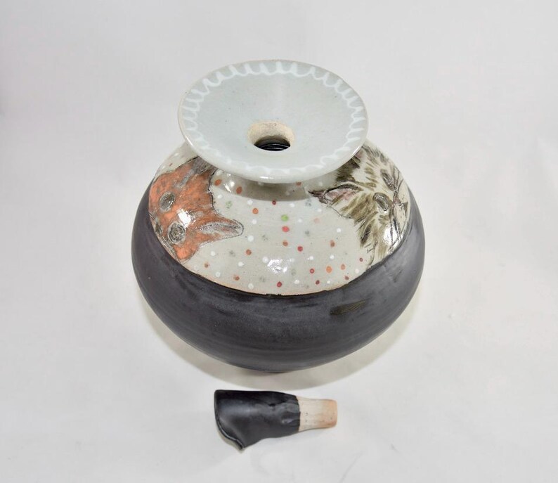 This is a handmade urn for holding a cat's ashes. It is ceramic and was made in the artist's Oregon pottery studio. This is a top view where you can see drawings of cats on the shoulder of the urn and the stopper laying beside it.