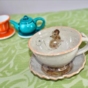 Puede incluir: Juego de taza y platillo de cer&aacute;mica con un dise&ntilde;o de rat&oacute;n pintado a mano. La taza es de color blanquecino con un borde naranja claro, y el platillo tiene un borde festoneado. Otras tazas y una tetera turquesa est&aacute;n en el fondo.