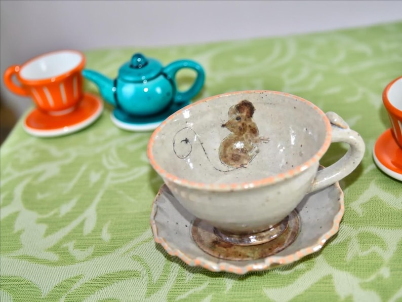Puede incluir: Juego de taza y platillo de cer&aacute;mica con un dise&ntilde;o de rat&oacute;n pintado a mano. La taza es de color blanquecino con un borde naranja claro, y el platillo tiene un borde festoneado. Otras tazas y una tetera turquesa est&aacute;n en el fondo.