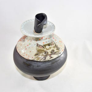 A ceramic cat-themed decorative pet urn with a black base and a cream-colored top. The jar features a hand-painted cat face and colorful polka dots. A black, curved stopper sits inside this urn to keep the ashes secure.
