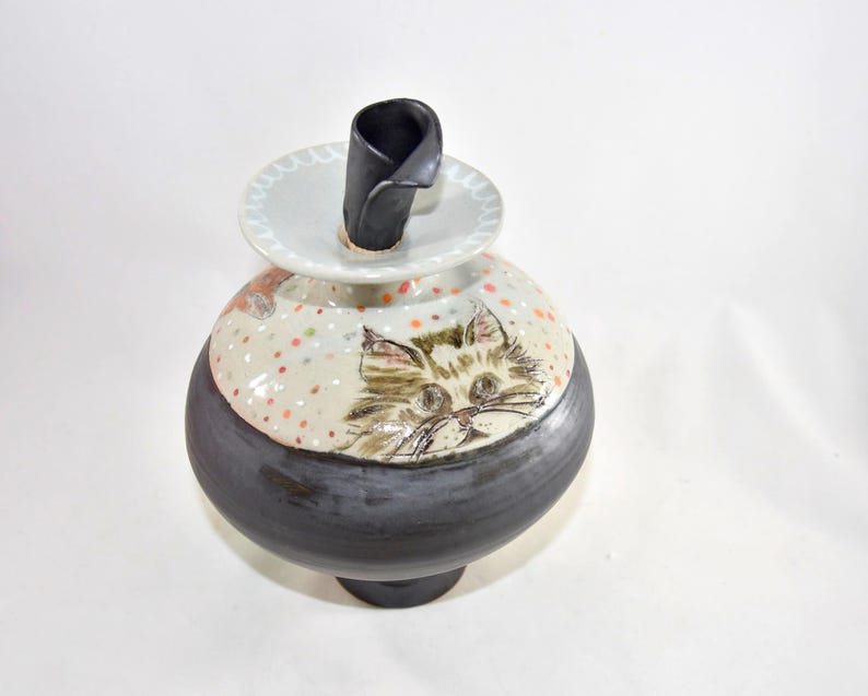 A ceramic cat-themed decorative pet urn with a black base and a cream-colored top. The jar features a hand-painted cat face and colorful polka dots. A black, curved stopper sits inside this urn to keep the ashes secure.