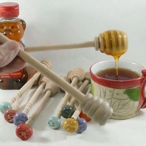 May include: A collection of wooden honey dippers with colorful ceramic handles featuring paw print designs. One dipper is held up, showing honey dripping into a ceramic mug filled with tea, while a bear-shaped honey bottle sits in the background.