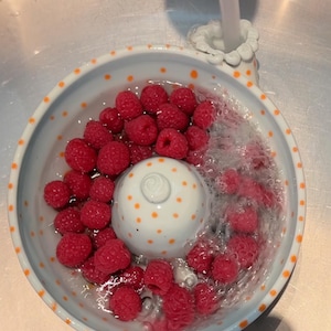May include: A white ceramic berry colander with orange polka dots, featuring a central raised dome and a side handle, filled with fresh red raspberries being rinsed under a stream of water from a faucet, creating bubbles in the water.