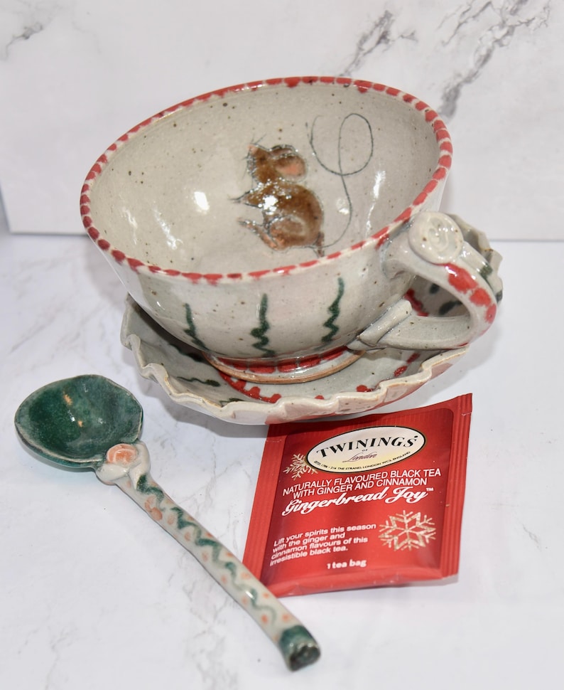 Puede incluir: Un juego de taza y platillo de cer&aacute;mica con un dise&ntilde;o de rat&oacute;n pintado a mano. El juego incluye una cuchara a juego y un paquete de t&eacute; Twinings Gingerbread Joy. La taza y el platillo tienen detalles en rojo y verde.