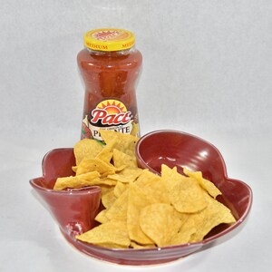 May include: A red ceramic chip and dip dish with a separate compartment for salsa. The dish is filled with tortilla chips and a jar of Pace Picante salsa is next to it.