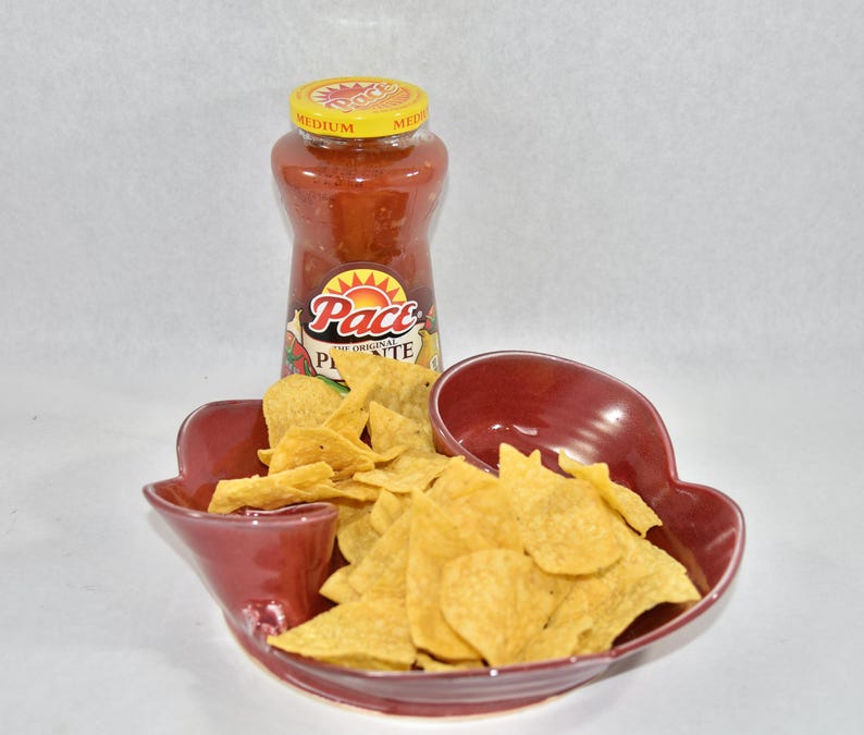 May include: A red ceramic chip and dip dish with a separate compartment for salsa. The dish is filled with tortilla chips and a jar of Pace Picante salsa is next to it.
