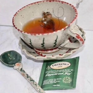 Puede incluir: Una taza de t&eacute; de cer&aacute;mica caprichosa con una figura de rat&oacute;n en su interior, llena de t&eacute;. La taza se encuentra sobre un platillo a juego, acompa&ntilde;ada de una cuchara de cer&aacute;mica verde y una bolsita de t&eacute; verde "Gingerbread Joy".