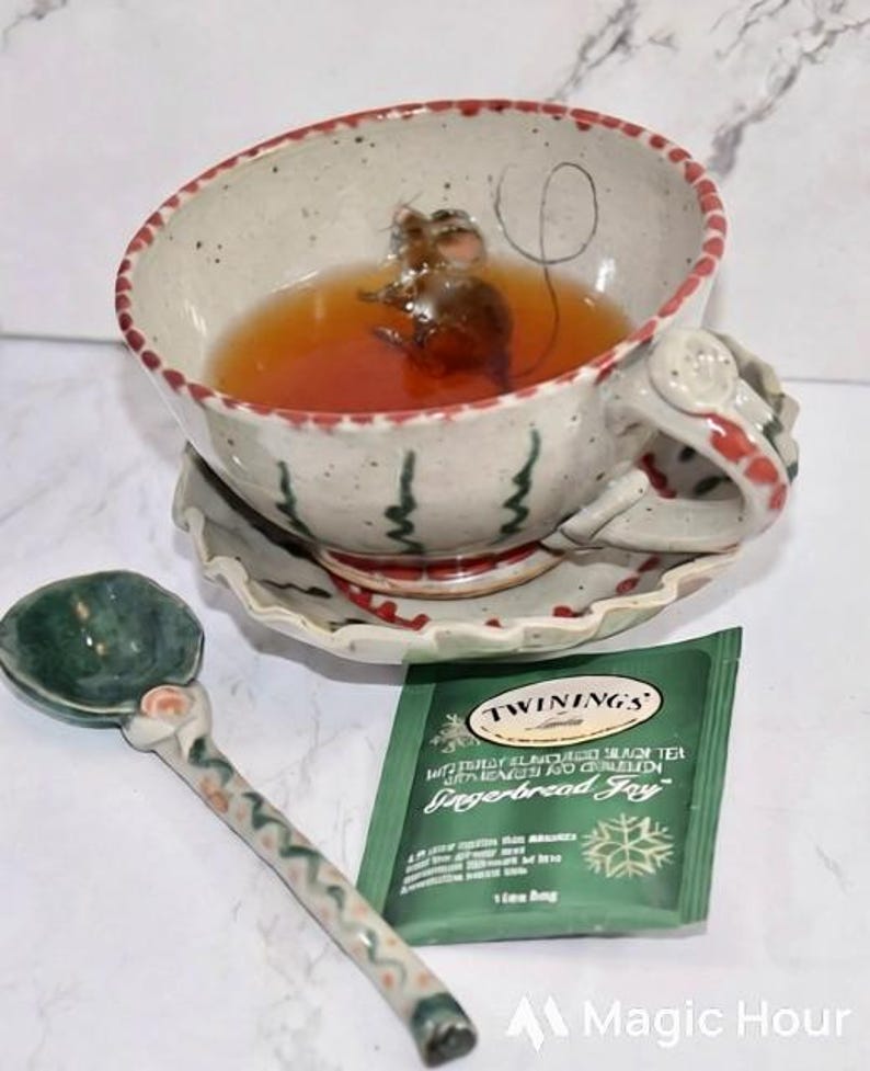 Puede incluir: Una taza de t&eacute; de cer&aacute;mica caprichosa con una figura de rat&oacute;n en su interior, llena de t&eacute;. La taza se encuentra sobre un platillo a juego, acompa&ntilde;ada de una cuchara de cer&aacute;mica verde y una bolsita de t&eacute; verde "Gingerbread Joy".