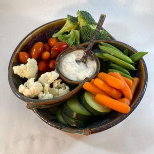 May include: A blue and brown ceramic serving dish with four sections. The dish is filled with a variety of vegetables, including broccoli, cauliflower, carrots, cucumbers, and green beans. A small white bowl in the center of the dish contains a white dip with a spoon.