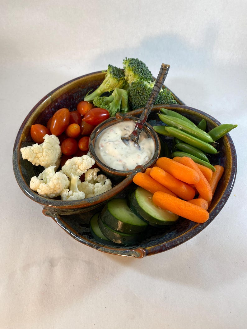 May include: A blue and brown ceramic serving dish with four sections. The dish is filled with a variety of vegetables, including broccoli, cauliflower, carrots, cucumbers, and green beans. A small white bowl in the center of the dish contains a white dip with a spoon.