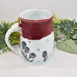 May include: A handmade ceramic mug with a burgundy glaze and a white floral design. The mug has a unique handle with gold accents.