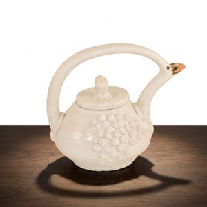 May include: A cream-colored ceramic teapot featuring a sculpted bird-shaped handle and spout, with a textured scale-like pattern on the body and a small bird-shaped knob on the lid. The spout tip is accented with a touch of brown color.