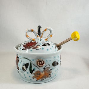 May include: A hand-painted ceramic sugar bowl with a lid and a wooden honey dipper. The bowl is light blue with a pattern of bees, leaves, and flowers. The lid has a bow-shaped handle and is decorated with polka dots.