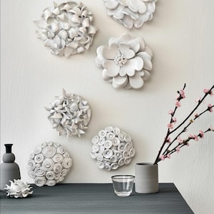 May include: A collection of six white ceramic wall sculptures shaped like various flowers and abstract circular patterns. The pieces are arranged on a wall above a dark gray table that holds a small gray vase with pink blossoms and a glass.