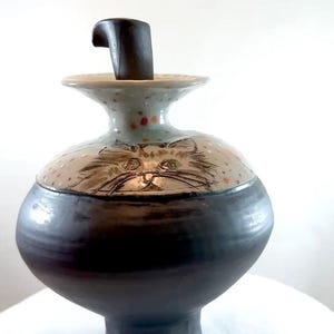 A 9.75 inch high ceramic urn for holding pet ashes and featuring drawings of two cats on the shoulder of this vessel. It has a unique shape with a deep foot and a stopper that fits into the top.