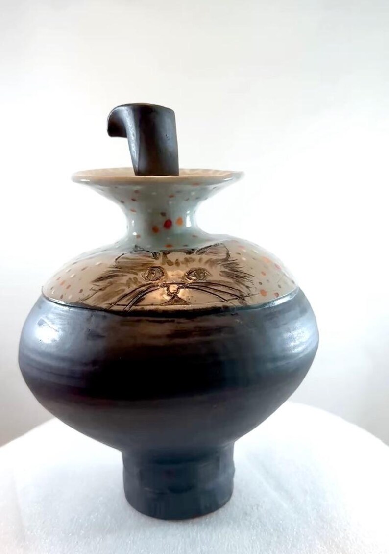 A 9.75 inch high ceramic urn for holding pet ashes and featuring drawings of two cats on the shoulder of this vessel. It has a unique shape with a deep foot and a stopper that fits into the top.