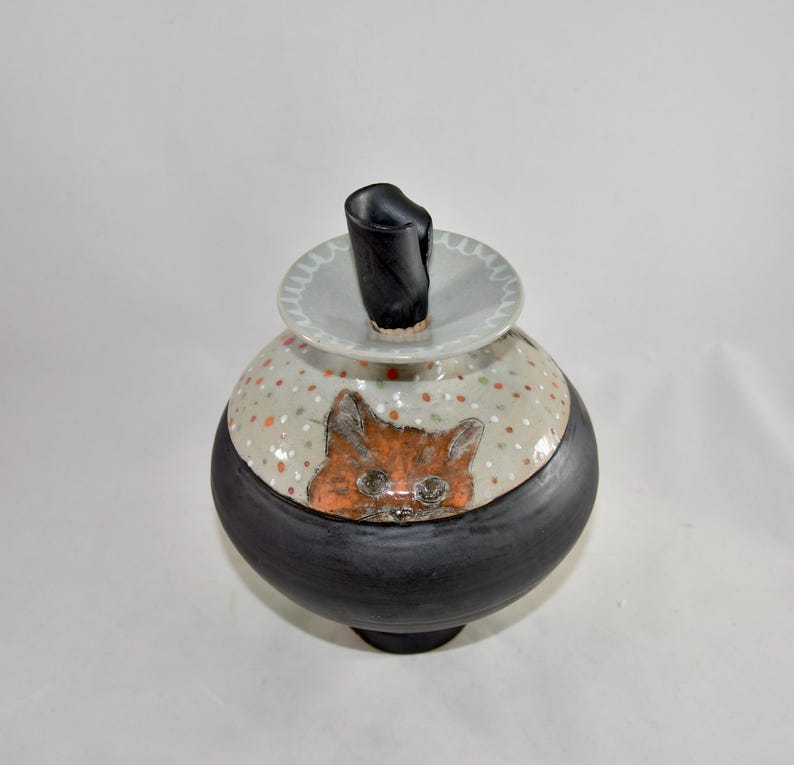 May include: A ceramic jar with a black base and a rounded body. The body is decorated with a cat face and polka dots. The lid has a black handle. The jar is a unique piece of art.