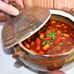 May include: A ceramic casserole dish with a lid, filled with a stew of vegetables and meat. The stew is a deep red color, with visible pieces of carrots, potatoes, and green onions. The dish has a brown and tan glaze.