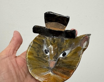 May include: A ceramic dish shaped like a cat head with a glossy finish. The cat is brown and tan with black eyes, a pink nose, and white whiskers. A black top hat is attached to the top of the cat head. The surface has a smooth, reflective glaze.