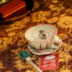 Puede incluir: Un juego de taza y platillo de cer&aacute;mica con un dise&ntilde;o de rat&oacute;n pintado a mano, una cuchara verde y blanca y una bolsita de t&eacute; roja sobre un mantel estampado. La taza tiene detalles rojos y verdes.