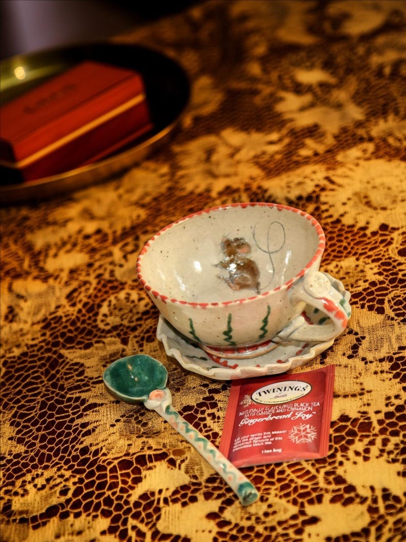 Puede incluir: Un juego de taza y platillo de cer&aacute;mica con un dise&ntilde;o de rat&oacute;n pintado a mano, una cuchara verde y blanca y una bolsita de t&eacute; roja sobre un mantel estampado. La taza tiene detalles rojos y verdes.