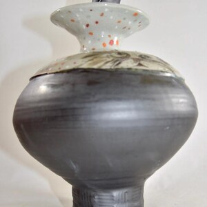 May include: A handcrafted ceramic vase with a dark gray body and a speckled lid. The lid features orange and white spots and a black handle. The vase has a bulbous shape and a textured base, showcasing a unique pottery design.
