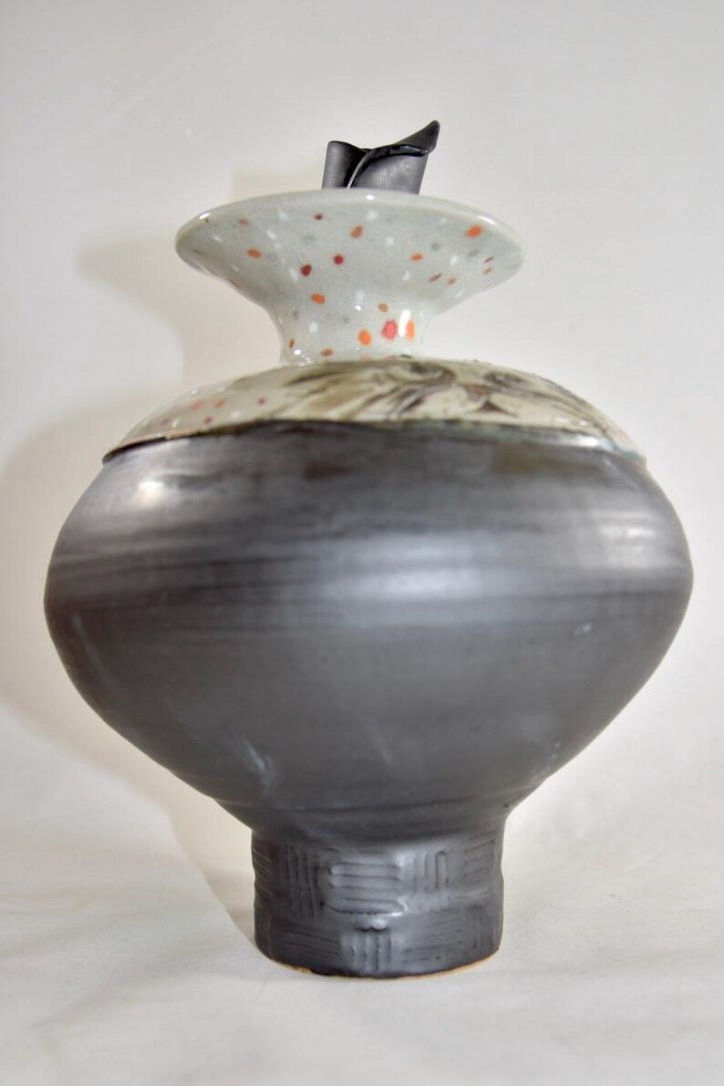 May include: A handcrafted ceramic vase with a dark gray body and a speckled lid. The lid features orange and white spots and a black handle. The vase has a bulbous shape and a textured base, showcasing a unique pottery design.