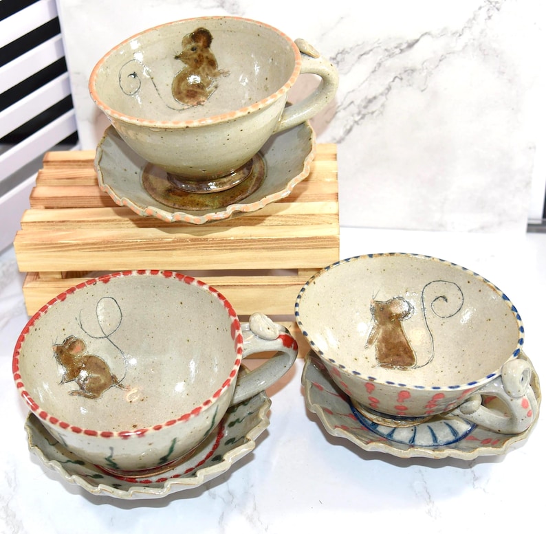 Puede incluir: Tazas de t&eacute; y platillos de cer&aacute;mica hechos a mano con ilustraciones de ratones. Las tazas tienen un color base beige con un dise&ntilde;o de rat&oacute;n y un platillo a juego. Los bordes est&aacute;n decorados con detalles en rojo, rosa y azul.
