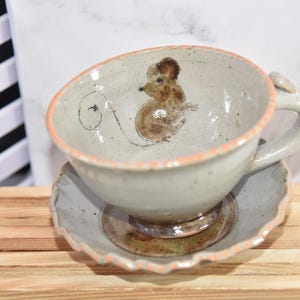Puede incluir: Un juego de taza y platillo de cer&aacute;mica. La taza presenta una ilustraci&oacute;n de rat&oacute;n pintada a mano en el interior. El borde de la taza y el platillo tienen un borde color melocot&oacute;n. El juego est&aacute; colocado sobre una superficie de madera.