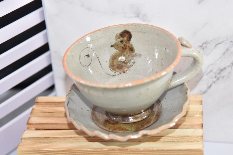 Puede incluir: Un juego de taza y platillo de cer&aacute;mica. La taza presenta una ilustraci&oacute;n de rat&oacute;n pintada a mano en el interior. El borde de la taza y el platillo tienen un borde color melocot&oacute;n. El juego est&aacute; colocado sobre una superficie de madera.