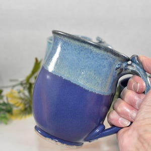 May include: A ceramic mug featuring a two-tone glaze with a deep royal blue base and a lighter, mottled sky blue upper section. The mug has a curved handle with decorative molded details at the top and bottom attachment points to the body.
