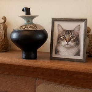 A pet urn sitting next to a photo of the deceased cat. This is a ceramic urn measuring 9.75 inches high and was made by the artist in her Oregon studio. It is glazed in a matte black on the bottom third and the stopper in the top. The top third features artwork of a gray cat and an orange cat.