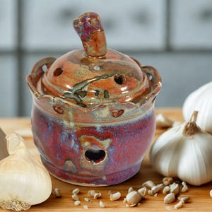 A handmade ceramic garlic keeper with a lid and knob that will hold around 3 garlic bulbs. The keeper is glazed in shades of red, brown, and blue. The lid and jar has several small holes for ventilation and a decorative edge.