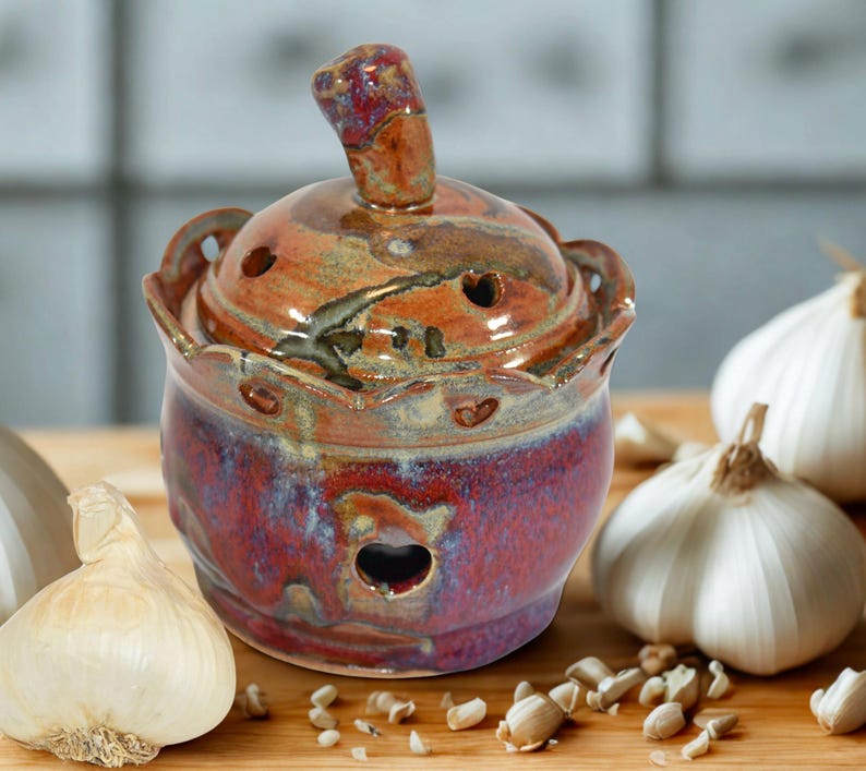 A handmade ceramic garlic keeper with a lid and knob that will hold around 3 garlic bulbs. The keeper is glazed in shades of red, brown, and blue. The lid and jar has several small holes for ventilation and a decorative edge.