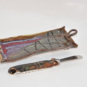 May include: A rectangular ceramic tray with two side handles featuring a mottled glaze in shades of brown, blue, and grey with dark dotted line patterns, accompanied by a matching spreader knife with a metal blade and a decorative ceramic handle.