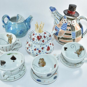 Puede incluir: Una colecci&oacute;n de tazas de t&eacute;, platillos y teteras de cer&aacute;mica pintadas a mano. Las tazas presentan ilustraciones de ratones y dise&ntilde;os de palos de cartas. Las teteras incluyen un dise&ntilde;o de cochecito de beb&eacute; azul, una corona y un dise&ntilde;o de patchwork con sombrero de copa.