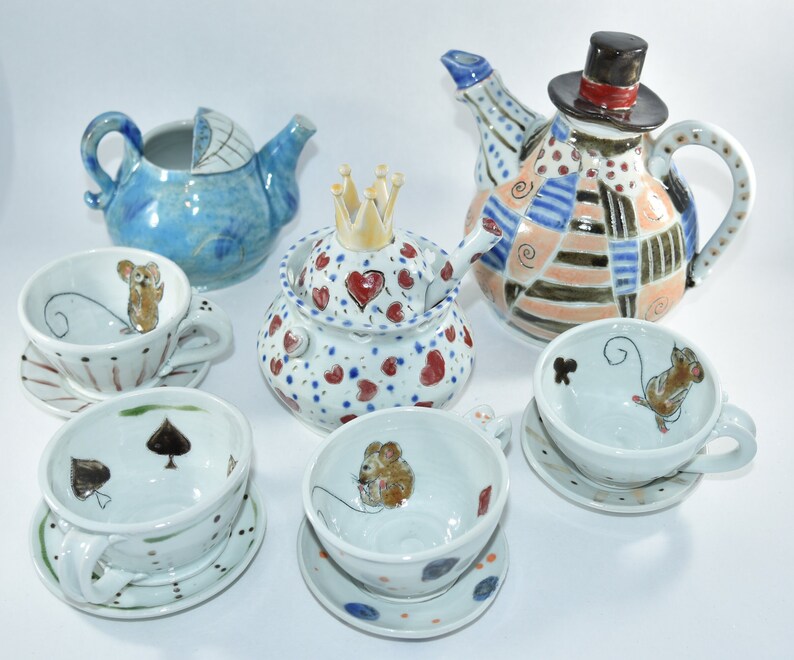 Puede incluir: Una colecci&oacute;n de tazas de t&eacute;, platillos y teteras de cer&aacute;mica pintadas a mano. Las tazas presentan ilustraciones de ratones y dise&ntilde;os de palos de cartas. Las teteras incluyen un dise&ntilde;o de cochecito de beb&eacute; azul, una corona y un dise&ntilde;o de patchwork con sombrero de copa.