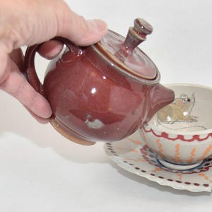 May include: A maroon ceramic teapot pouring into a matching teacup and saucer set. The teacup features a whimsical frog design inside. The saucer and teacup have a decorative border. The teapot is held by a person.