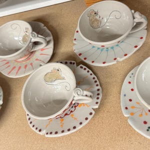 Puede incluir: Tazas y platillos de cer&aacute;mica blanca con ilustraciones de ratones. Las tazas tienen un asa curva y se asientan sobre platillos decorativos con patrones coloridos. El rat&oacute;n est&aacute; dibujado en la taza y el platillo.