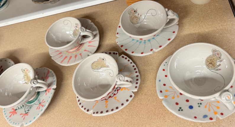 Puede incluir: Tazas y platillos de cer&aacute;mica blanca con ilustraciones de ratones. Las tazas tienen un asa curva y se asientan sobre platillos decorativos con patrones coloridos. El rat&oacute;n est&aacute; dibujado en la taza y el platillo.