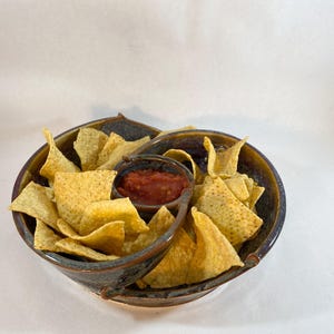 May include: A brown and blue ceramic chip and dip serving dish with a small bowl for salsa and a larger bowl for chips. The dish has two handles on the sides. The dish is filled with tortilla chips and salsa.