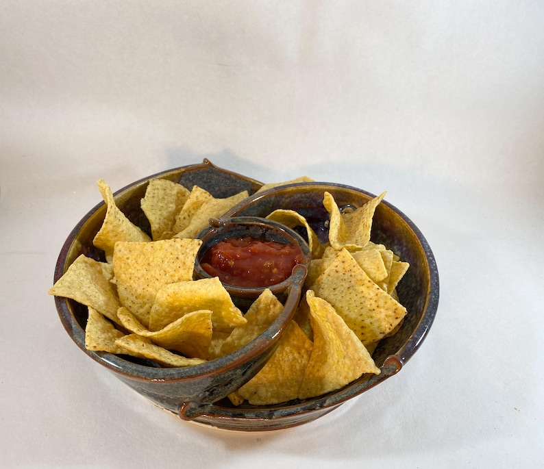 May include: A brown and blue ceramic chip and dip serving dish with a small bowl for salsa and a larger bowl for chips. The dish has two handles on the sides. The dish is filled with tortilla chips and salsa.
