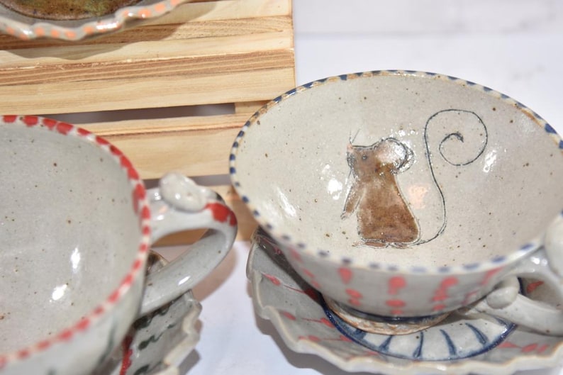 Puede incluir: Tazas de t&eacute; y platillos de cer&aacute;mica hechos a mano. Las tazas tienen un acabado moteado blanquecino con borde y asa rojos. Una taza presenta una ilustraci&oacute;n de rat&oacute;n marr&oacute;n. Los platillos tienen un borde festoneado con detalles rojos y azules.