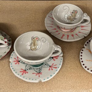 Puede incluir: Una colecci&oacute;n de tazas y platillos de cer&aacute;mica hechos a mano. Cada taza presenta una ilustraci&oacute;n caprichosa de un rat&oacute;n y se combina con un platillo decorado de forma &uacute;nica. Los platillos tienen patrones y dise&ntilde;os coloridos.