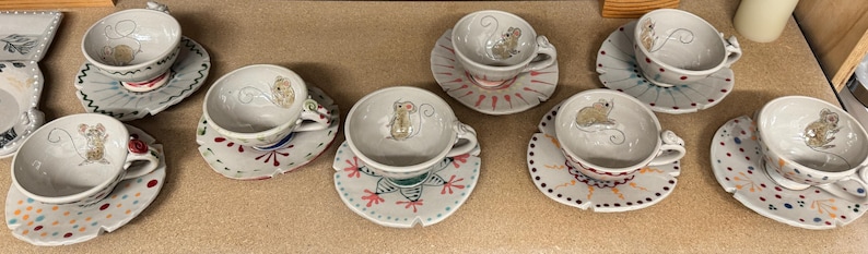 Puede incluir: Una colecci&oacute;n de tazas y platillos de cer&aacute;mica hechos a mano. Cada taza presenta una ilustraci&oacute;n caprichosa de un rat&oacute;n y se combina con un platillo decorado de forma &uacute;nica. Los platillos tienen patrones y dise&ntilde;os coloridos.