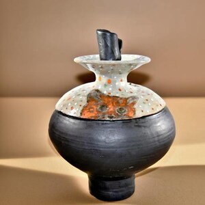 A ceramic urn with a black base and body, a white and gray dotted lid, and a black stopper that fits inside the opening. The top of the urn features an orange cat face design. The jar is a unique piece of home decor.