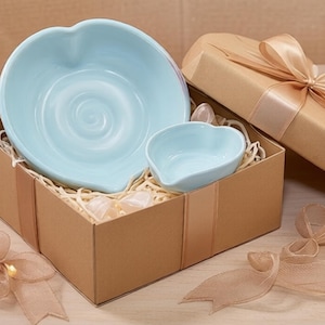 May include: A light blue ceramic bowl and a smaller matching light blue ceramic dish, both shaped like hearts, nestled inside a brown cardboard gift box filled with shredded paper filler and topped with a tan satin ribbon bow.